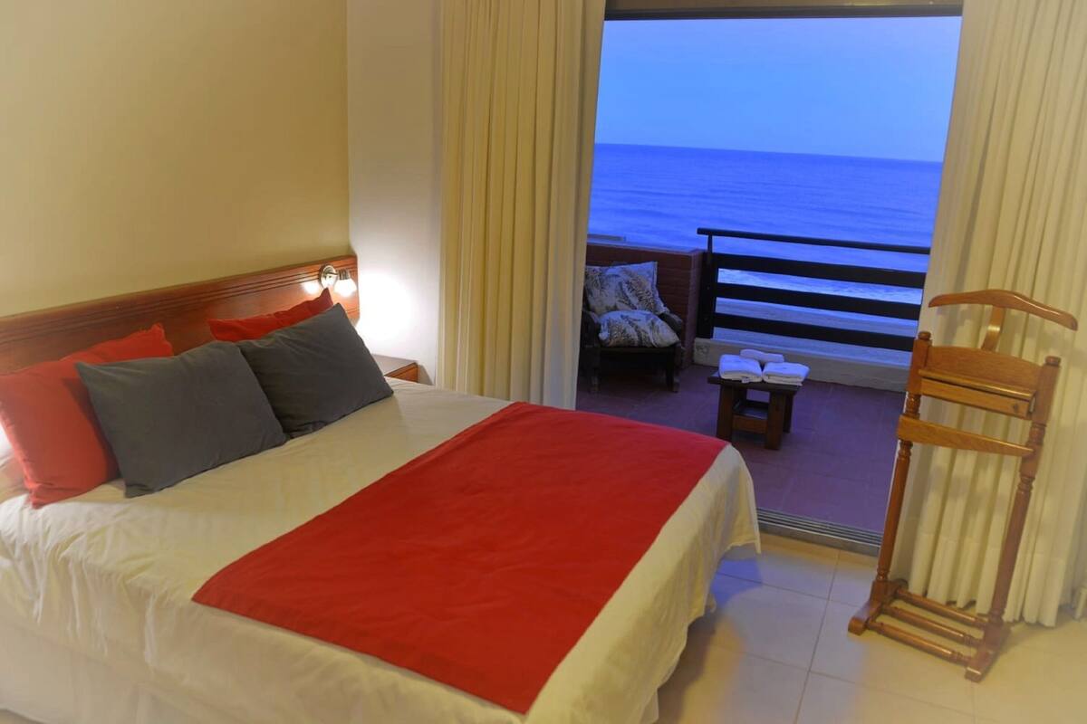 A hotel room with a bed, a wooden chair, a small table, and a view of the ocean through a balcony door.