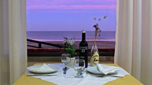 A table set with a bottle of wine, glasses, and a vase with flowers, overlooking a beach with a pink sky. - Intersur Villa Gesell (Villa Gesell)