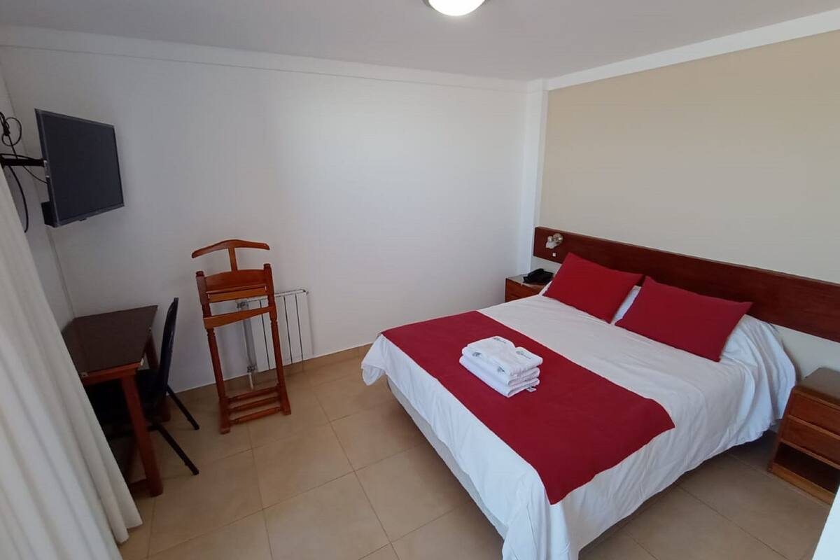 A hotel room with a bed, a desk, a chair, a TV, and a radiator.