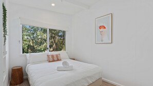 Een slaapkamer met een groot raam, een bed met wit beddengoed, een ingelijst schilderij aan de muur en een klein houten krukje.