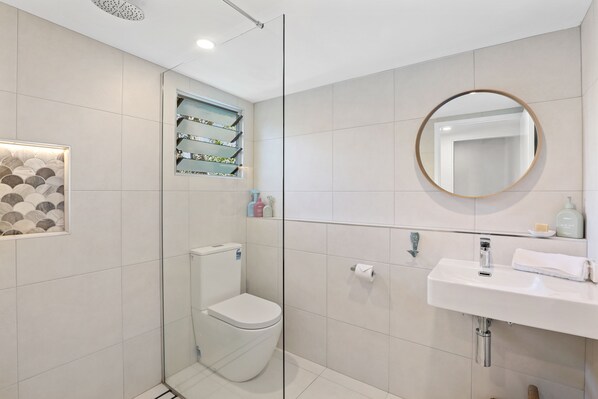 A modern bathroom with a glass shower enclosure, a toilet, a sink with a mirror, and a shelf with toiletries.