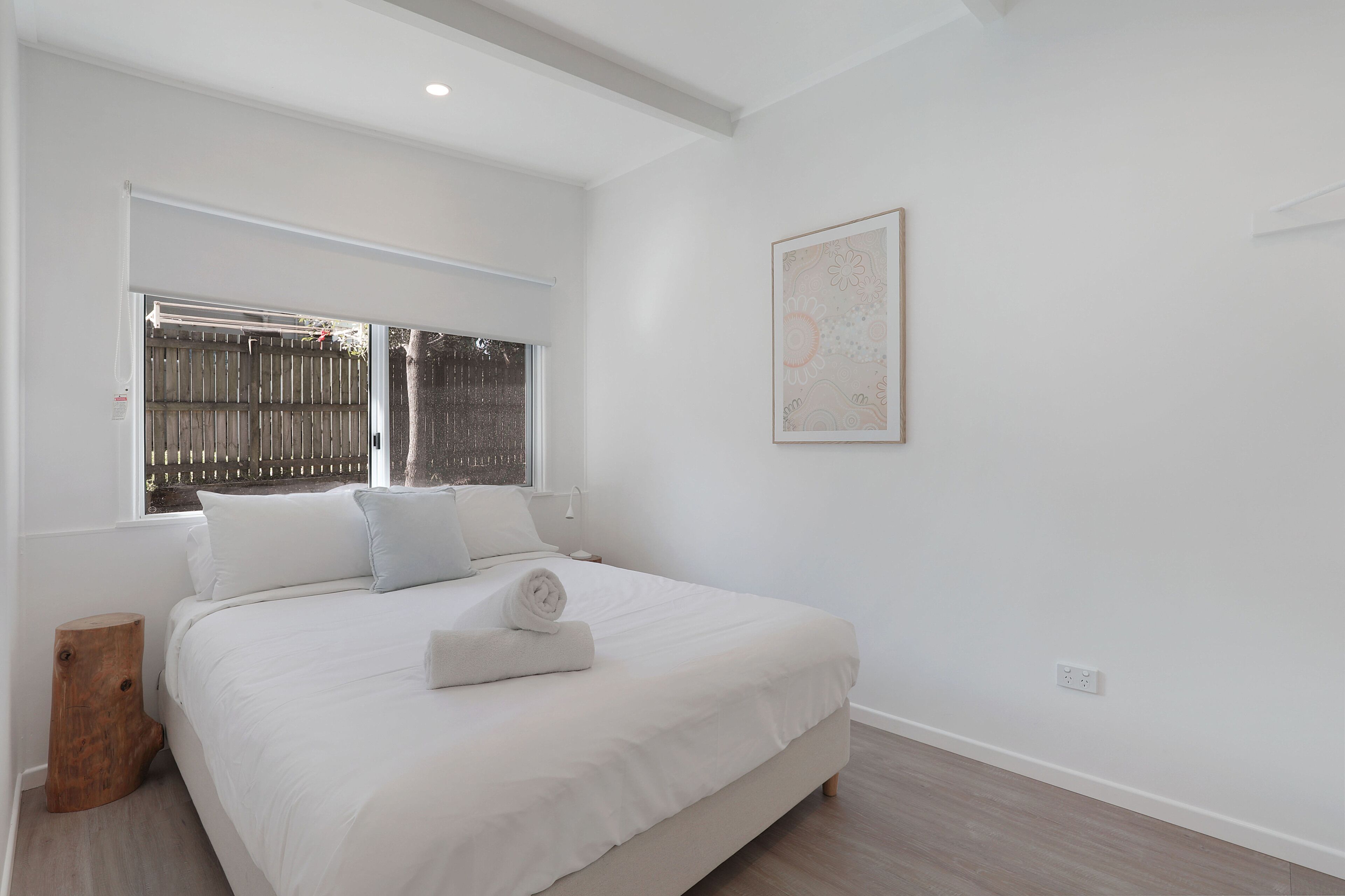 A bedroom with a large bed, a window with a view of a fence, a wall-mounted artwork, and a small wooden stool.