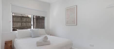 A bedroom with a large bed, a window with a view of a fence, a wall-mounted artwork, and a small wooden stool.