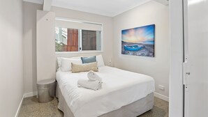 A neatly made bed with white linens and a rolled towel, a window with curtains, a painting of a sunset over water, and a trash bin. - 5 Unit Property - Lots of space and privacy for the whole group (Point Lookout)