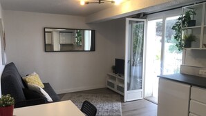 En moderne stue med sofa, spisebord og fjernsyn. En skydedør i glas fører ud til en balkon med planter.