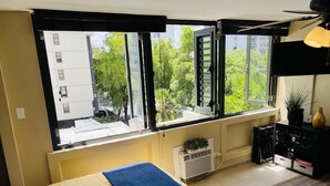 Une chambre à coucher avec un lit, un radiateur, une fenêtre donnant sur des immeubles et des arbres, et un ventilateur de plafond.
