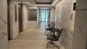 Un couloir moderne aux murs de marbre, un espace détente avec des chaises en métal et un ascenseur.