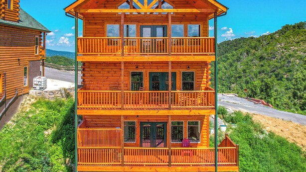 A multi-story wooden cabin with balconies, a stone foundation, and a view of a road and forested hills. - Free Attractions | Indoor Heated Pool, Game Room, Fireplace, Views (Sevierville)
