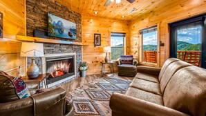 A cozy log cabin interior with a fireplace, leather seating, a television, and a scenic view through the windows. - Free Attractions | Indoor Heated Pool, Game Room, Fireplace, Views (Sevierville)