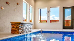 A swimming pool with a waterfall feature, a stone ledge, and a view of the sky through windows. - Free Attractions | Indoor Heated Pool, Game Room, Fireplace, Views (Sevierville)