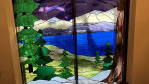 Ein Buntglasfenster mit einer Landschaftsdarstellung, darunter Berge, Bäume und ein Gewässer.