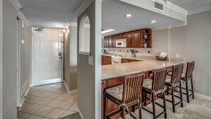 A kitchen with a central island, wooden bar stools, and a white door leading to another room. - 🌟Oceanfront | Awesome Amenities | Great Views | Carolina Dunes 204🌟 (Myrtle Beach)