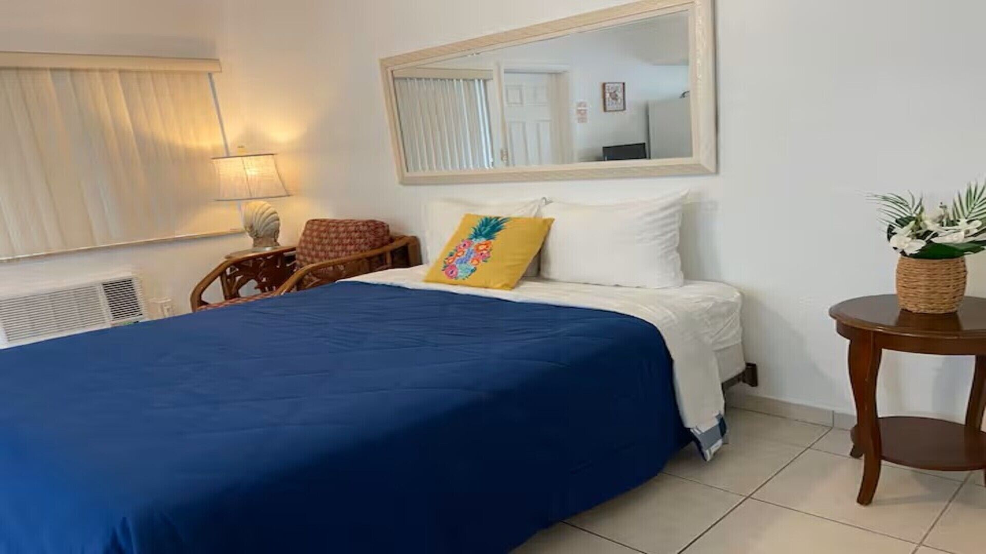 A hotel room with a bed, a mirror, a chair, a lamp, and a small table with a plant.