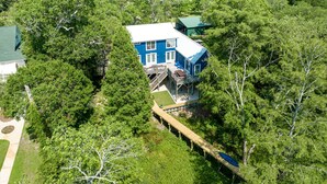 A blue house surrounded by dense forest, with a wooden deck and stairs leading to a porch.