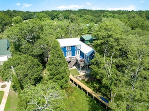 Una casa azul rodeada de un bosque frondoso, con una terraza de madera y escaleras que conducen a un porche.