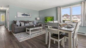 Una sala de estar moderna con un sofá gris, una mesa de centro y un comedor con sillas blancas y una mesa dispuesta para cuatro.