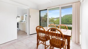 A dining area with a table and chairs, a kitchen in the background, and a view of greenery through a large window. - Tyrone Beach Escape (Rye)