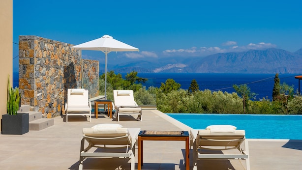 Un espace aménagé au bord de la piscine, avec des chaises longues et une table, d’où l’on peut admirer une mer d’un bleu limpide et des montagnes.