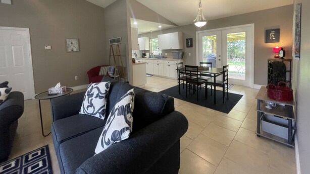 A living room with a sofa, a dining table with chairs, a kitchen in the background, and a staircase leading to another level.