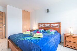 Una cama bien hecha con colcha azul y almohadas estampadas, un cabecero de madera y una mesita de noche con lámpara.