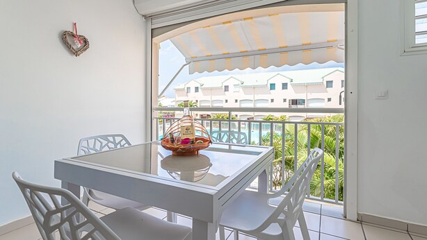 A balcony with a white table and chairs, a wicker basket with a tag, and a view of buildings and a pool. - Ô Rêves Bleus - Two Bedroom Apartment, Sleeps 4 (Saint-Francois)