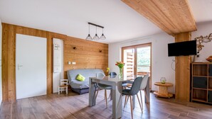 Una sala de estar moderna con techo de madera, una mesa de comedor con sillas, un sofá, un televisor montado en la pared y un ventanal que proporciona luz natural.