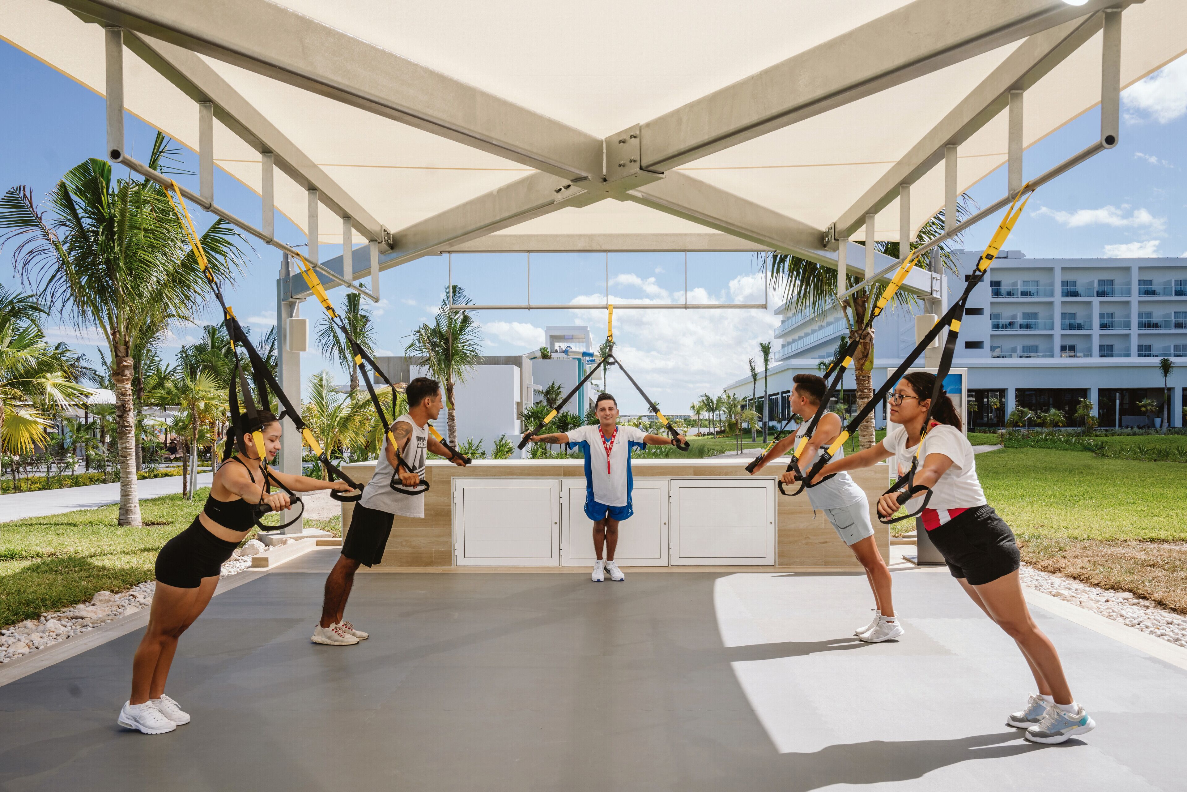 A group of people exercising with TRX suspension straps in an outdoor area with a covered structure, palm trees, and a building in the background.