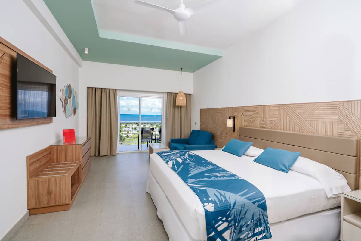 A hotel room with a bed, a TV, a desk, a chair, a balcony with a view, and a ceiling fan.
