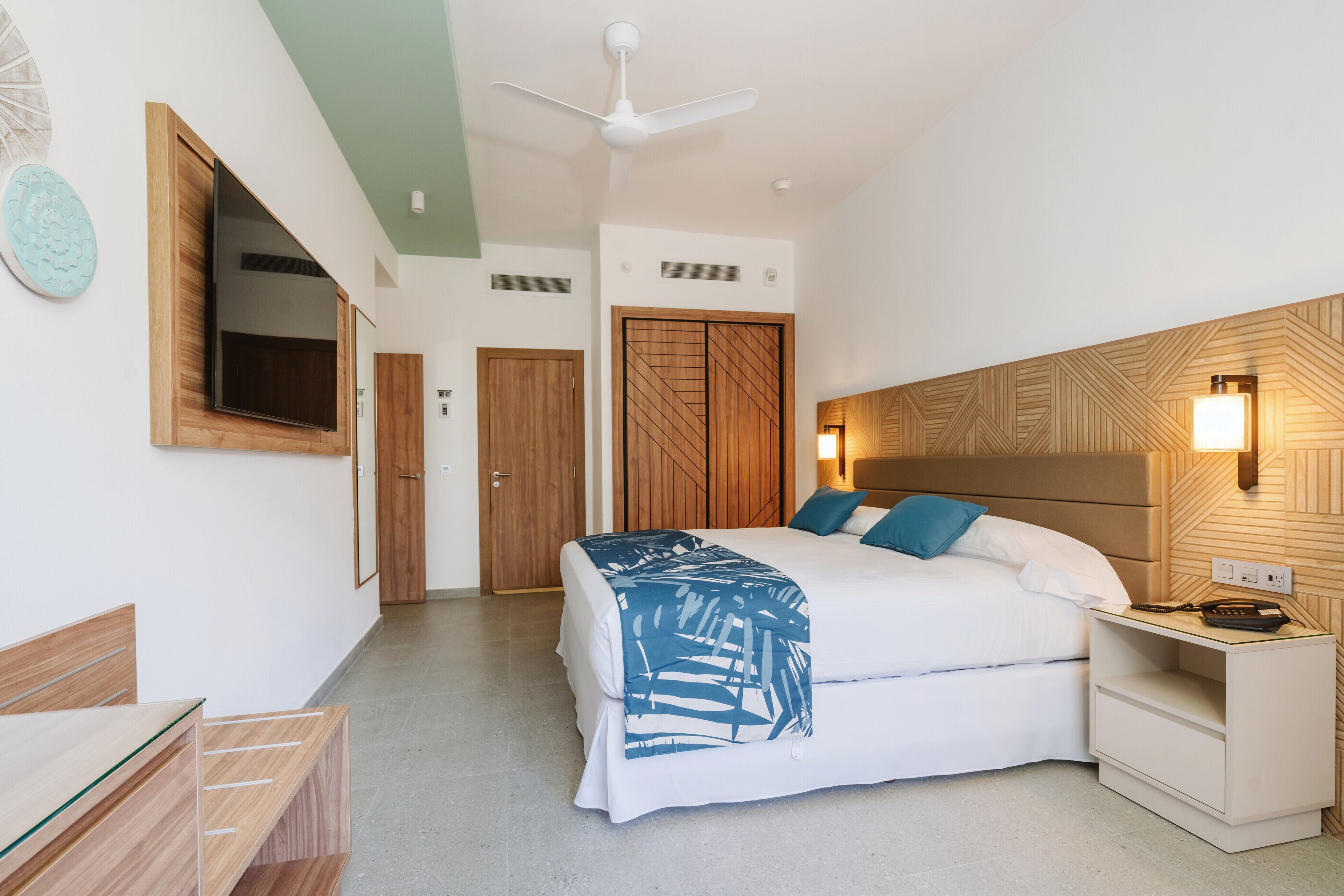 A hotel room with a bed, bedside table, nightstand, ceiling fan, and a wall-mounted TV.