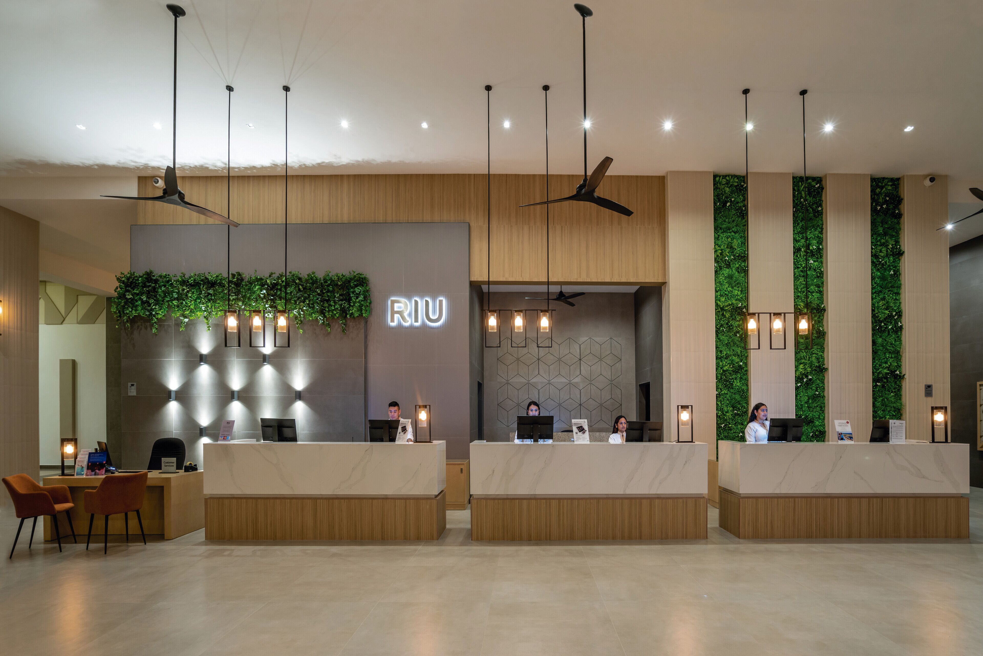 Un área de recepción moderna con un mostrador de madera, un jardín vertical verde y varios ventiladores de techo.