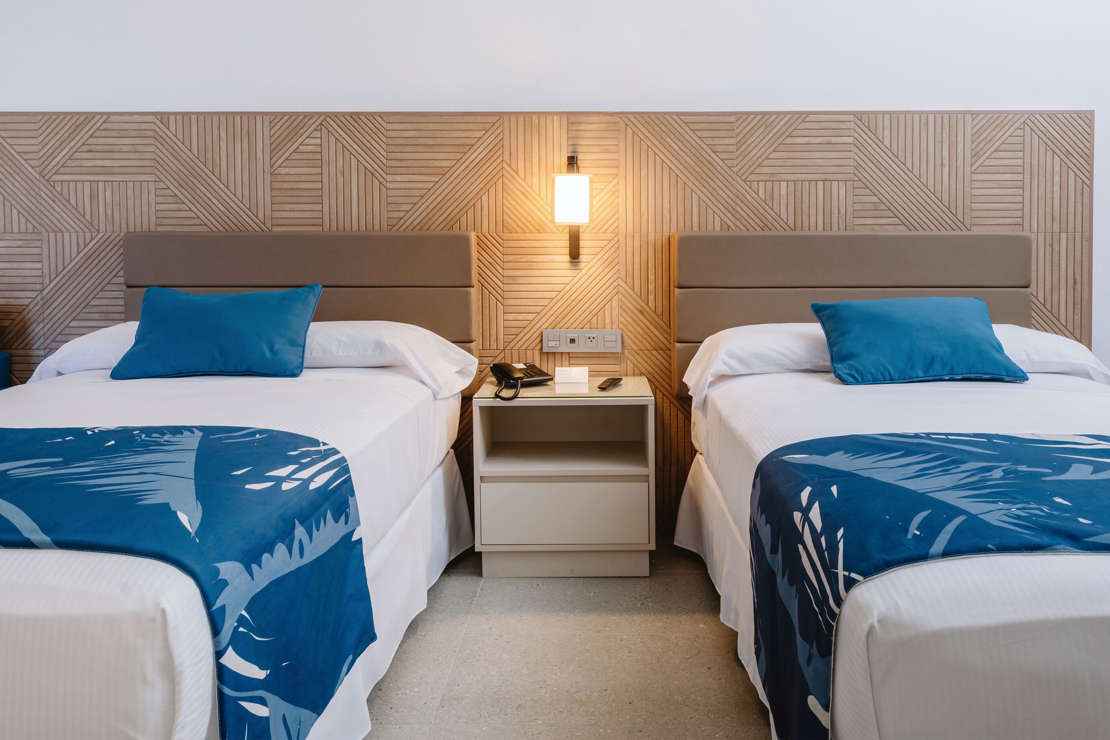 A hotel room with two beds, each with a blue bedspread and pillow. There is a nightstand between the beds with a lamp and a phone. The wall has a geometric pattern.