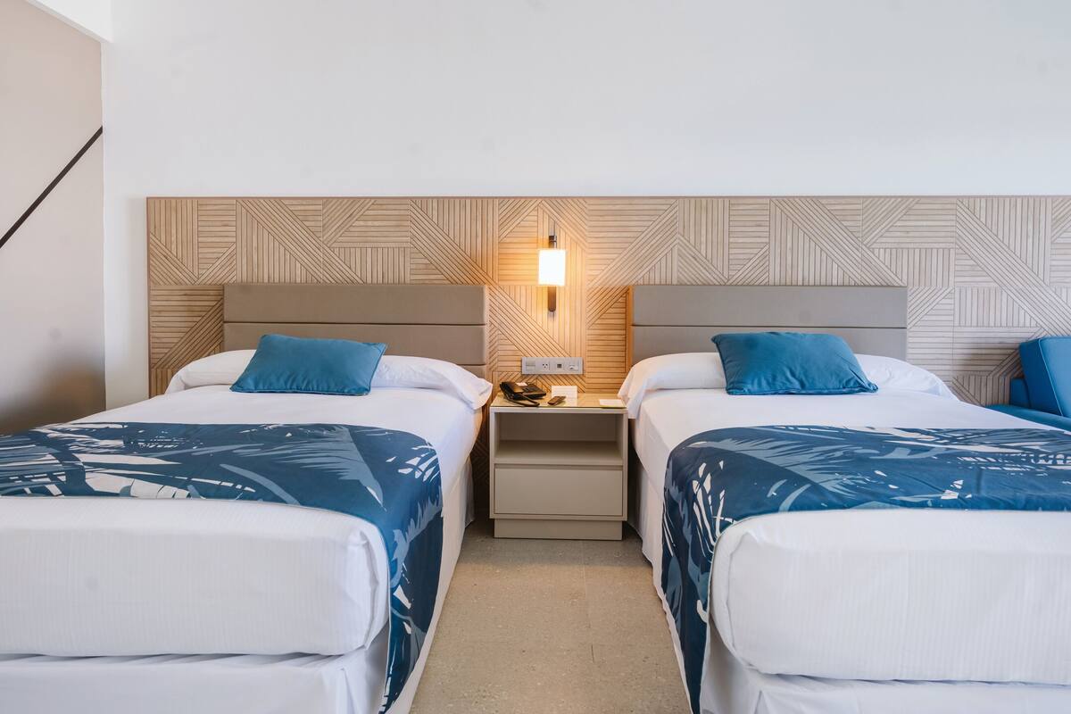 A hotel room with two beds, each with a headboard and a bedside table with a lamp.