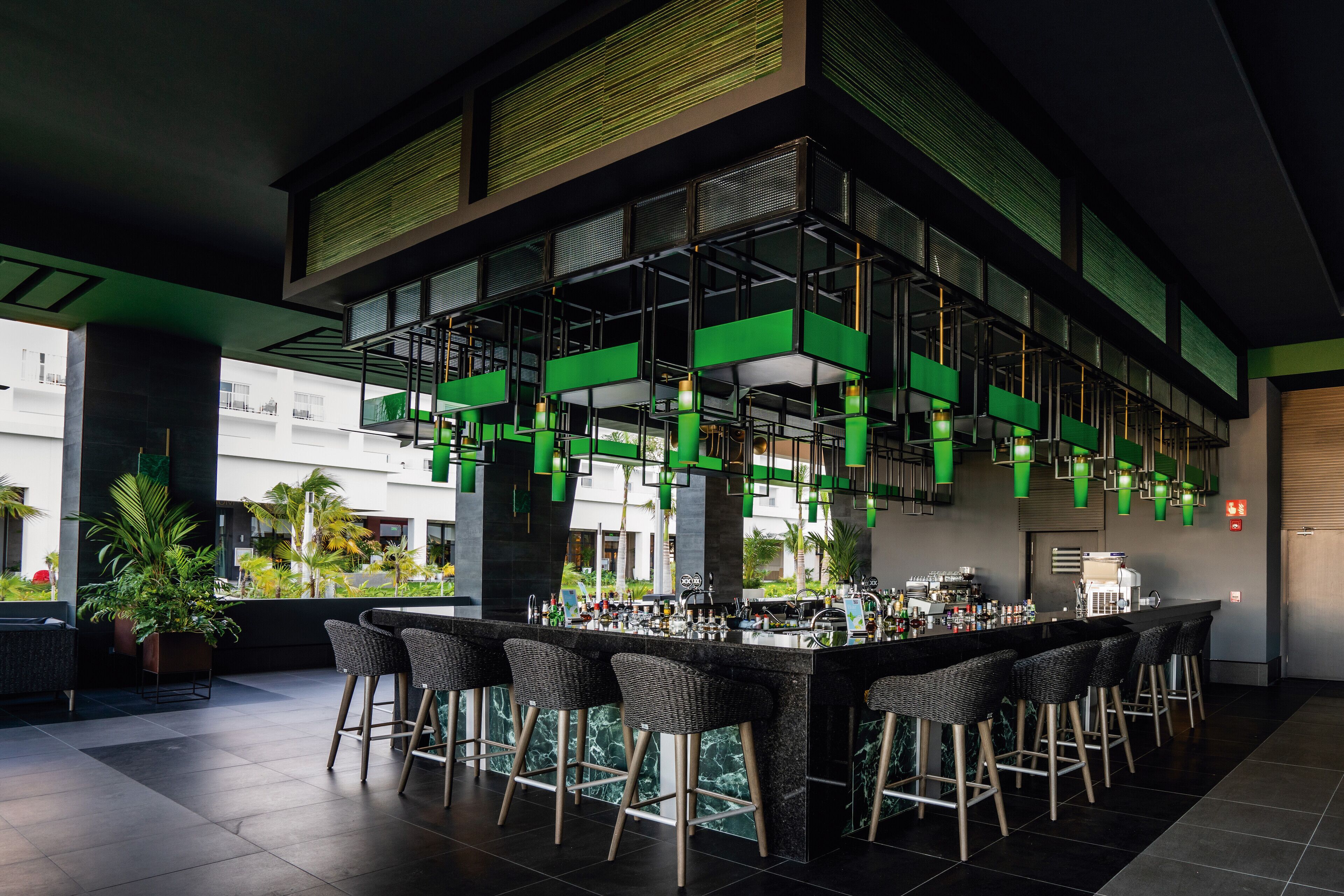 A modern bar area with a high bar counter, multiple stools, and a view of the outdoor area with plants.