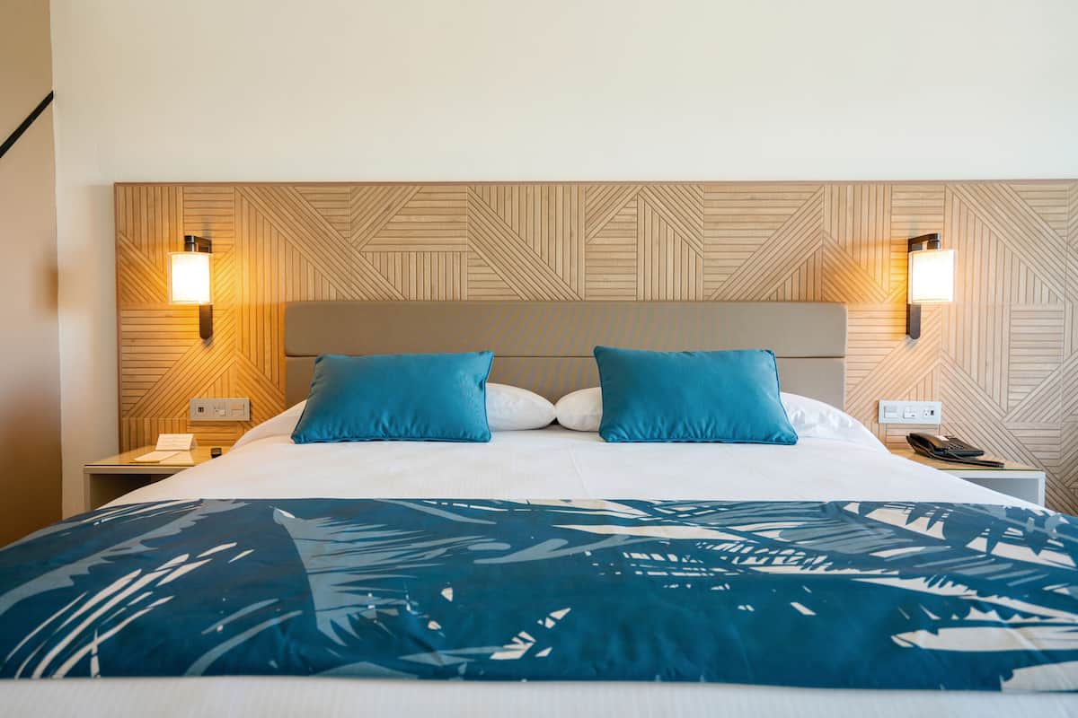 A hotel room with a bed, two blue pillows, a patterned headboard, and bedside lamps.