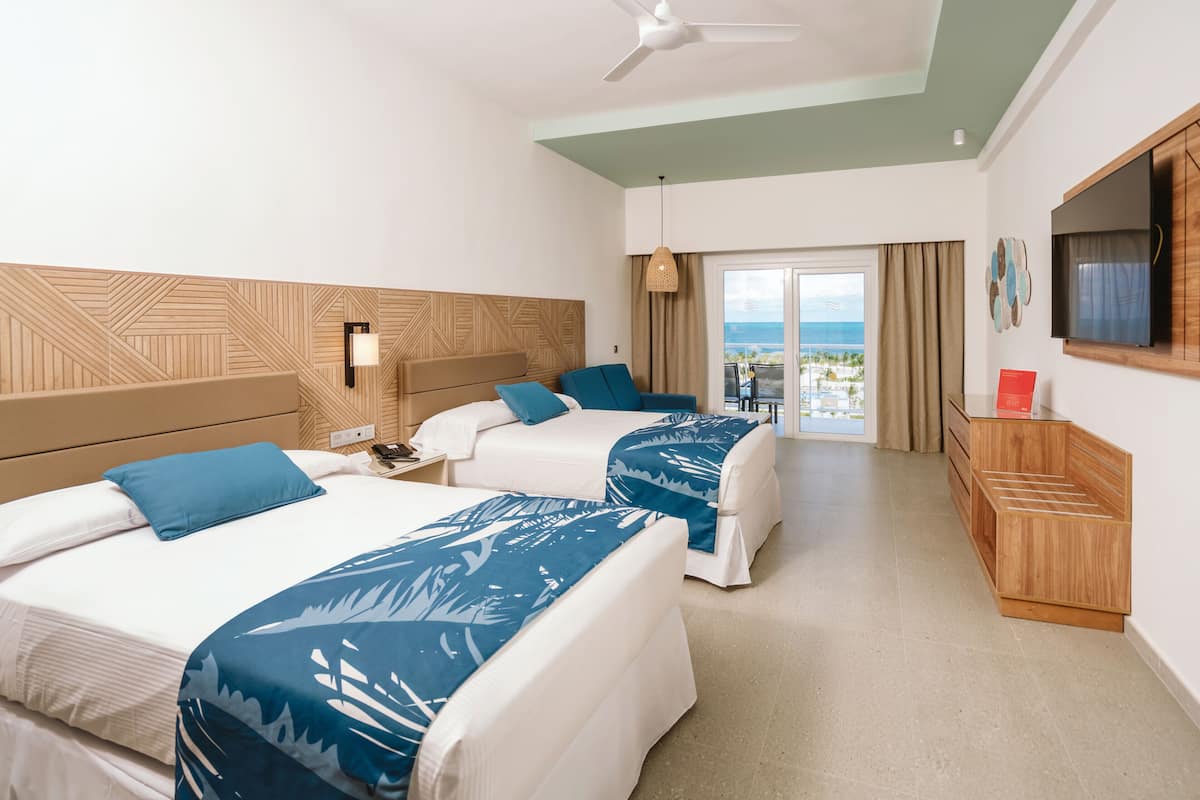A hotel room with two beds, a TV, a desk, and a balcony with a view of the sea.
