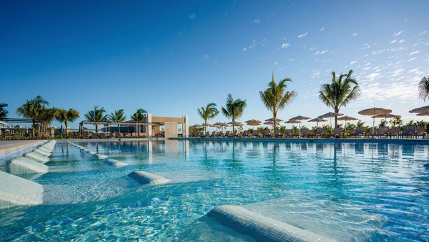 Una piscina azul cristalina con escalones revestidos de azulejos que bajan al agua, rodeada de palmeras y sillones de descanso bajo sombrillas.
