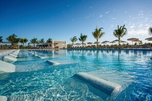 A clear blue pool with tiled steps leading into it, surrounded by palm trees and lounge chairs under umbrellas.