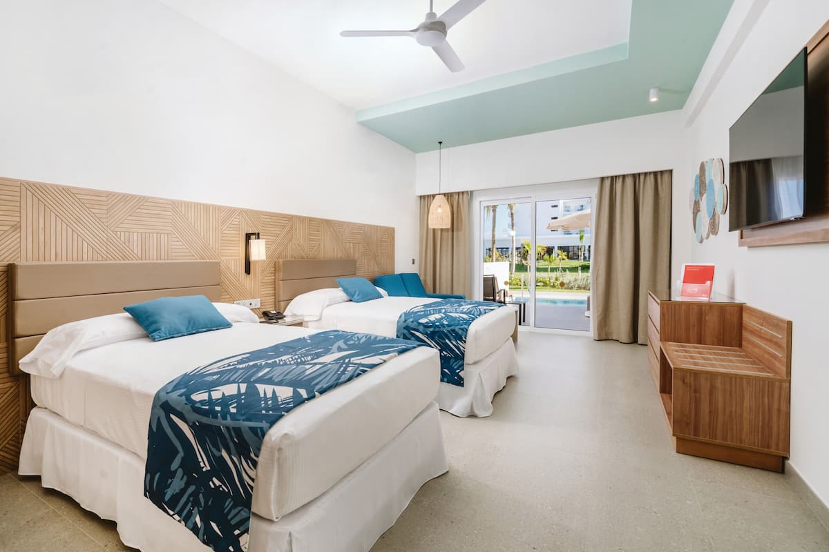 A hotel room with two beds, a ceiling fan, a TV, and a view of the pool.