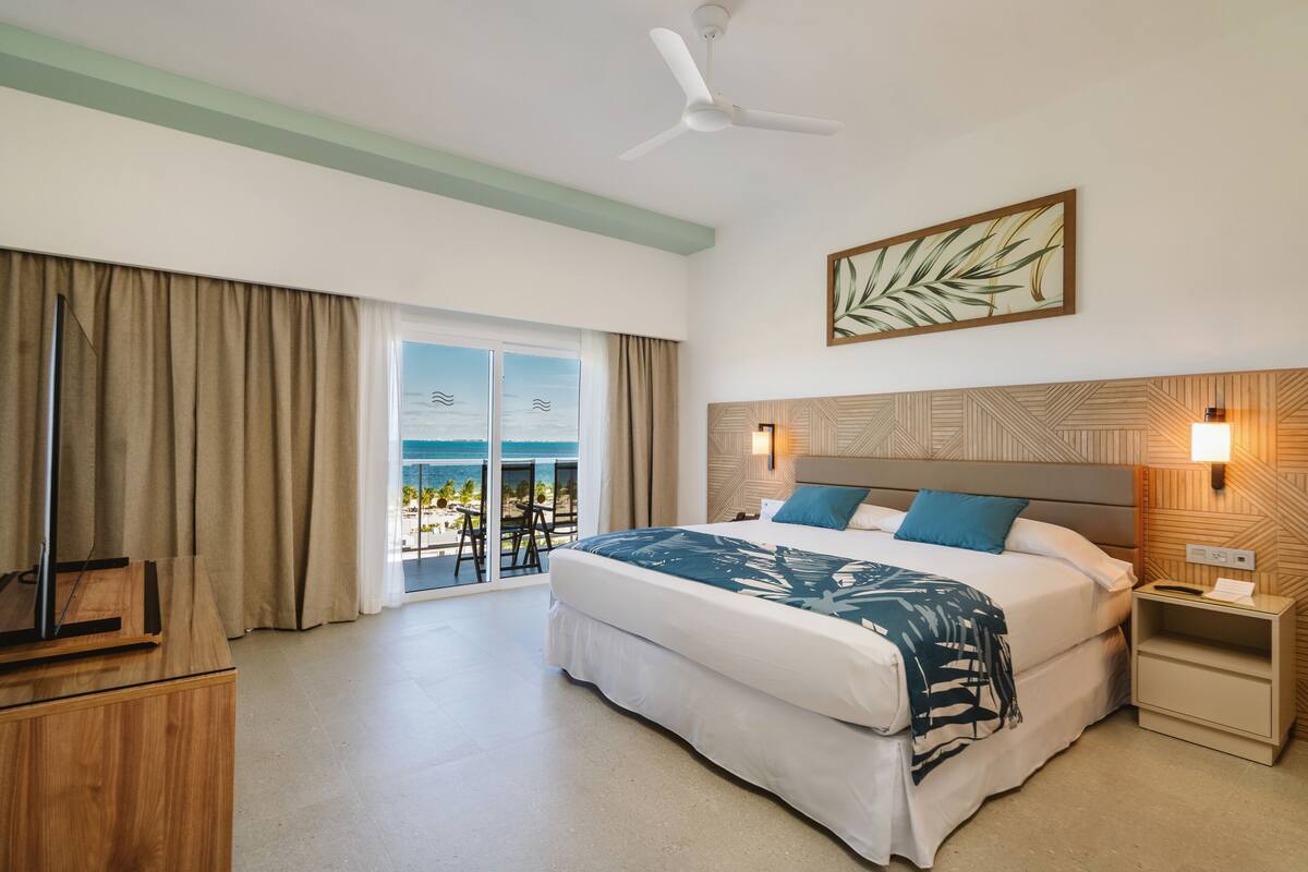 A hotel room with a bed, a ceiling fan, a nightstand, a desk, and a view of the ocean through a sliding door.