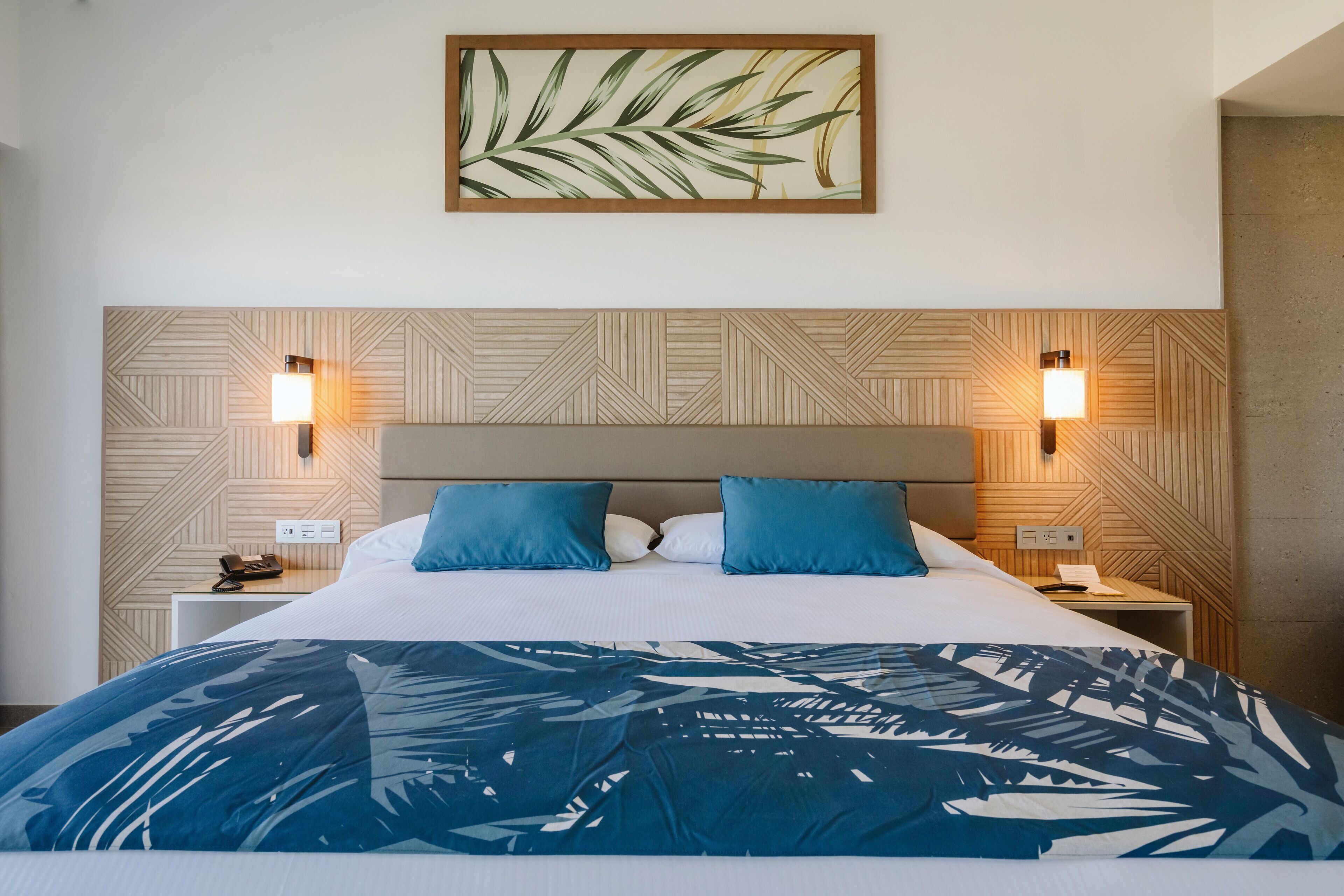 A hotel room with a bed, two bedside tables, a wall-mounted light fixture, and a framed artwork above the bed.