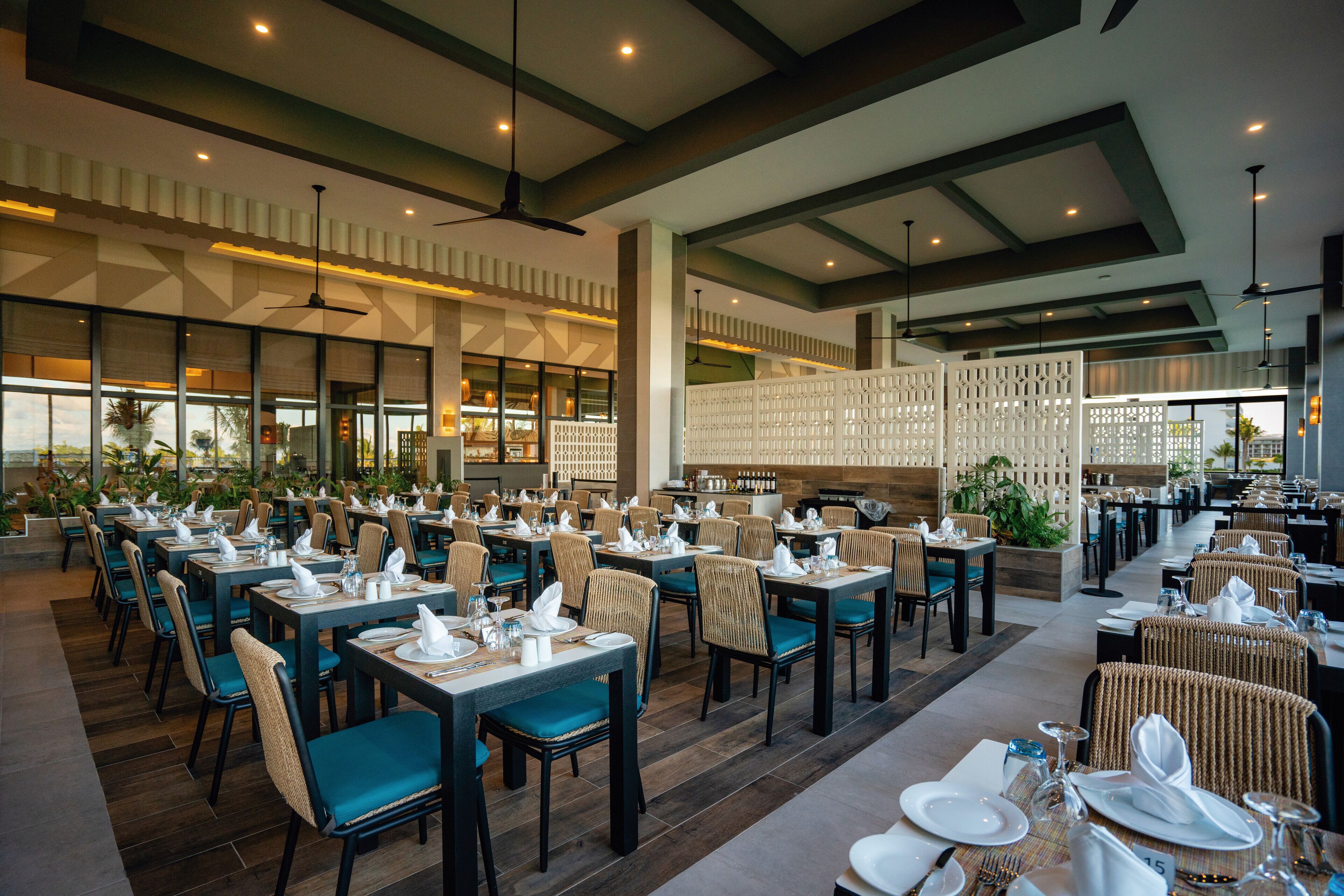Un'ampia sala ristorante con numerosi tavoli e sedie, soffitti alti con travi a vista e grandi finestre che offrono una vista.