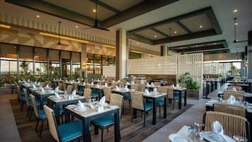 A spacious restaurant with multiple dining tables and chairs, high ceilings with exposed beams, and large windows offering views.