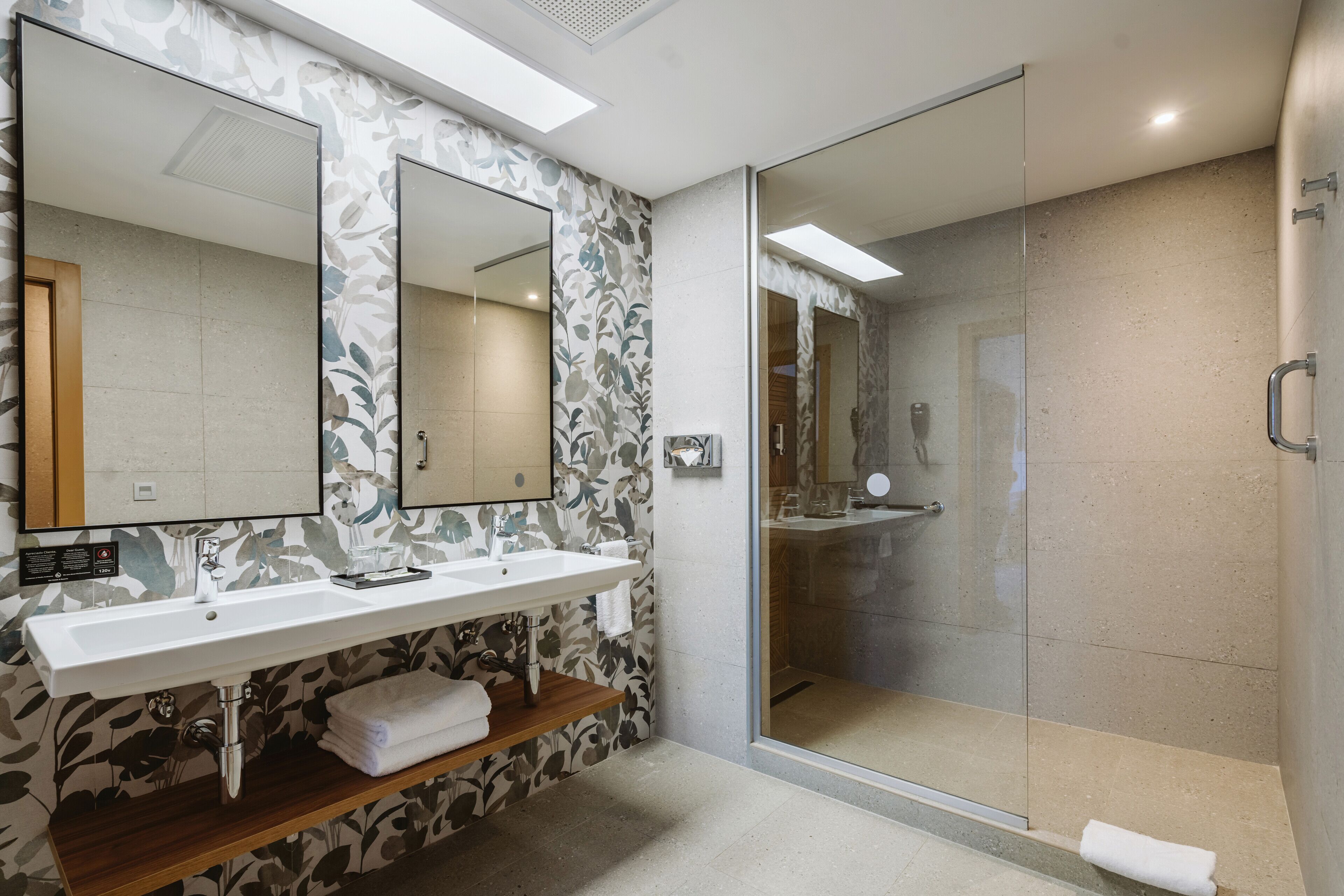 Un baño moderno con un espejo grande, dos lavamanos, una ducha y un estante de madera.