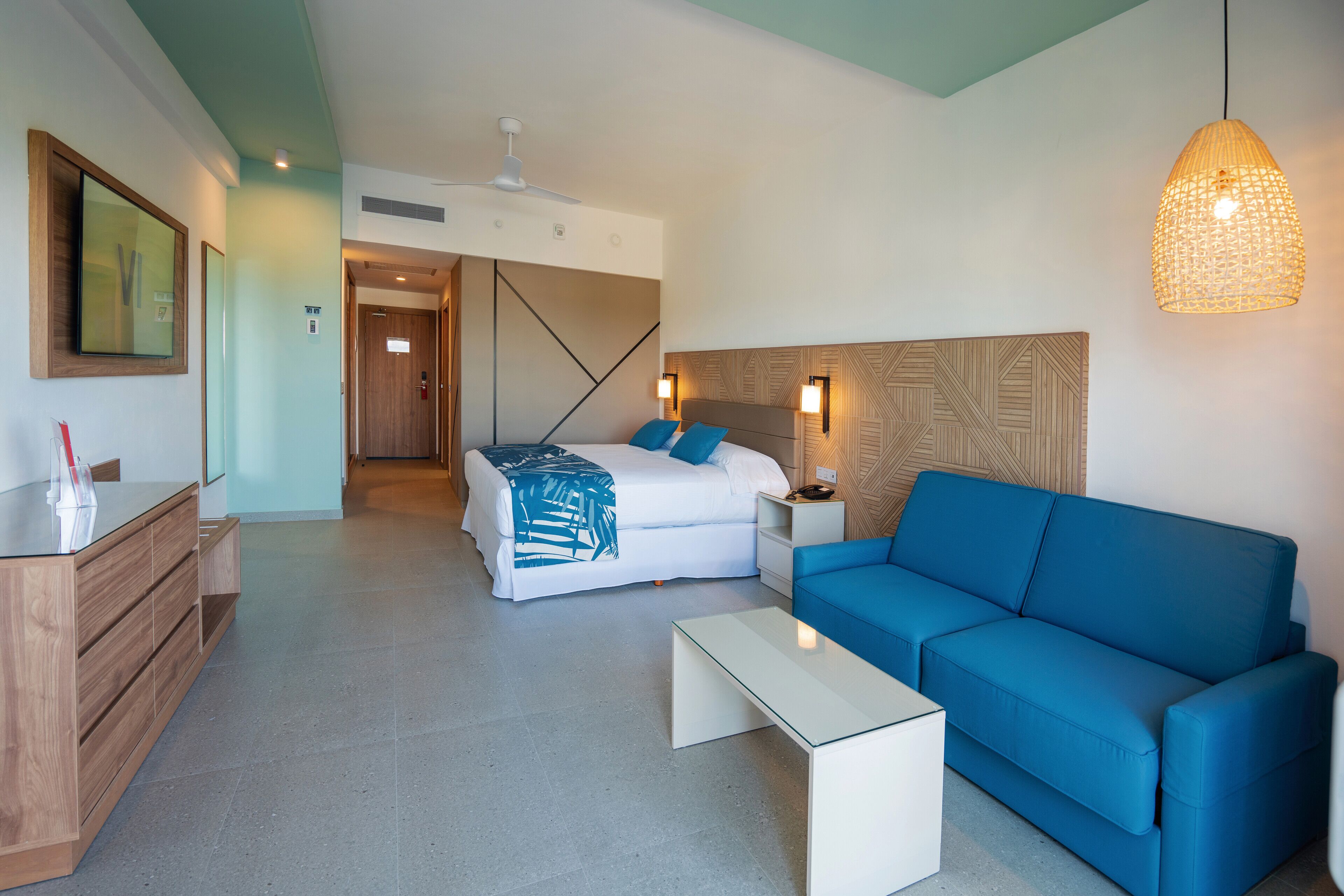 A modern hotel room with a bed, a blue sofa, a white coffee table, and a wooden dresser.