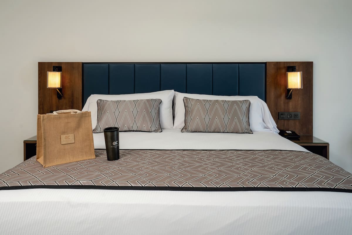A hotel room with a large bed, two pillows, a nightstand, a lamp, a bag, and a cup.