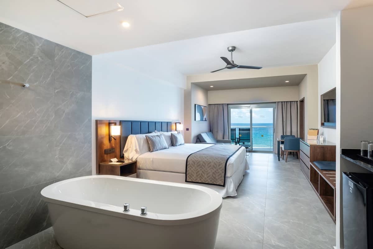 A modern hotel room with a large bed, a bathtub, a desk, and a view of the sea.