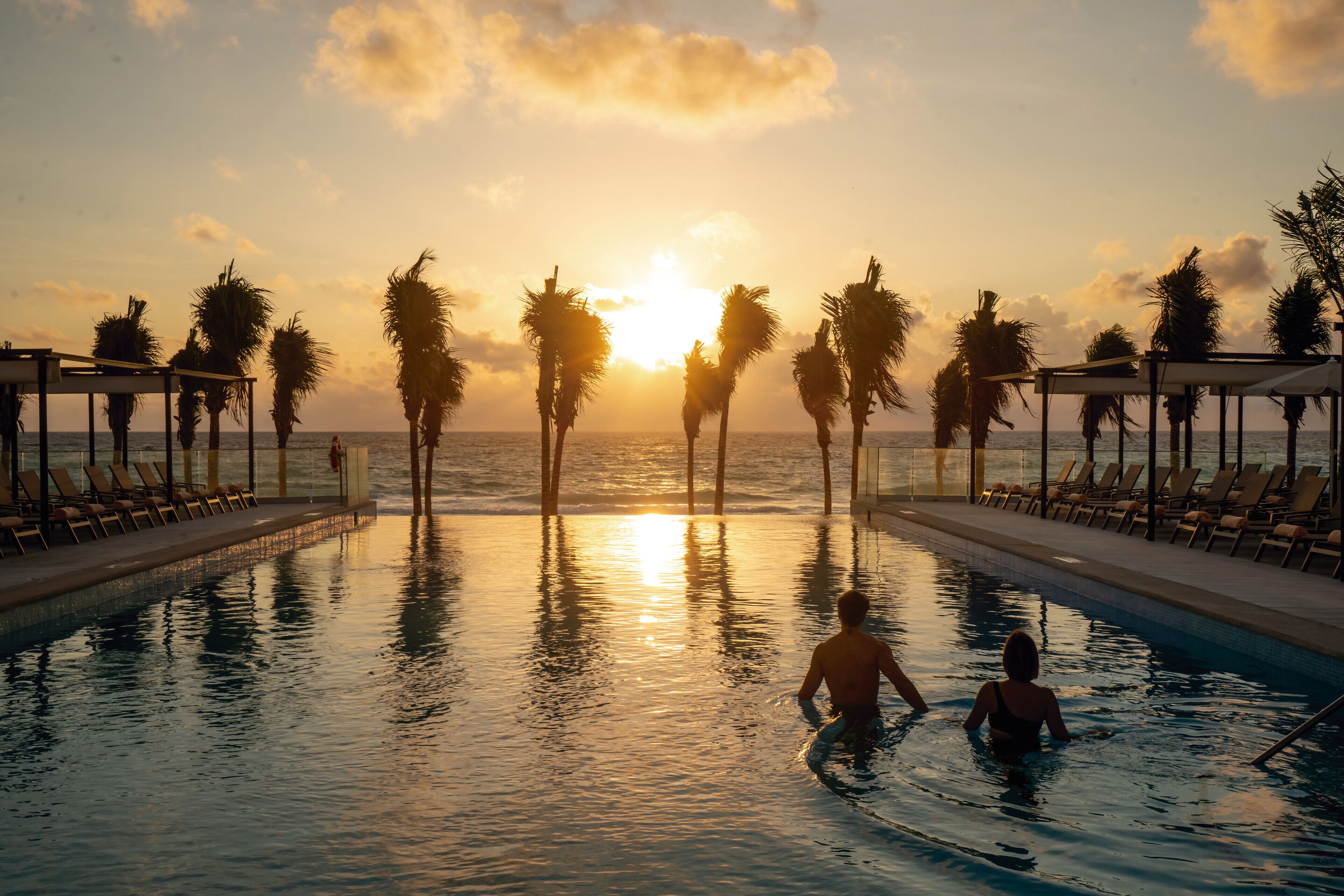 A swimming pool with lounge chairs and palm trees, with the sun setting in the background.