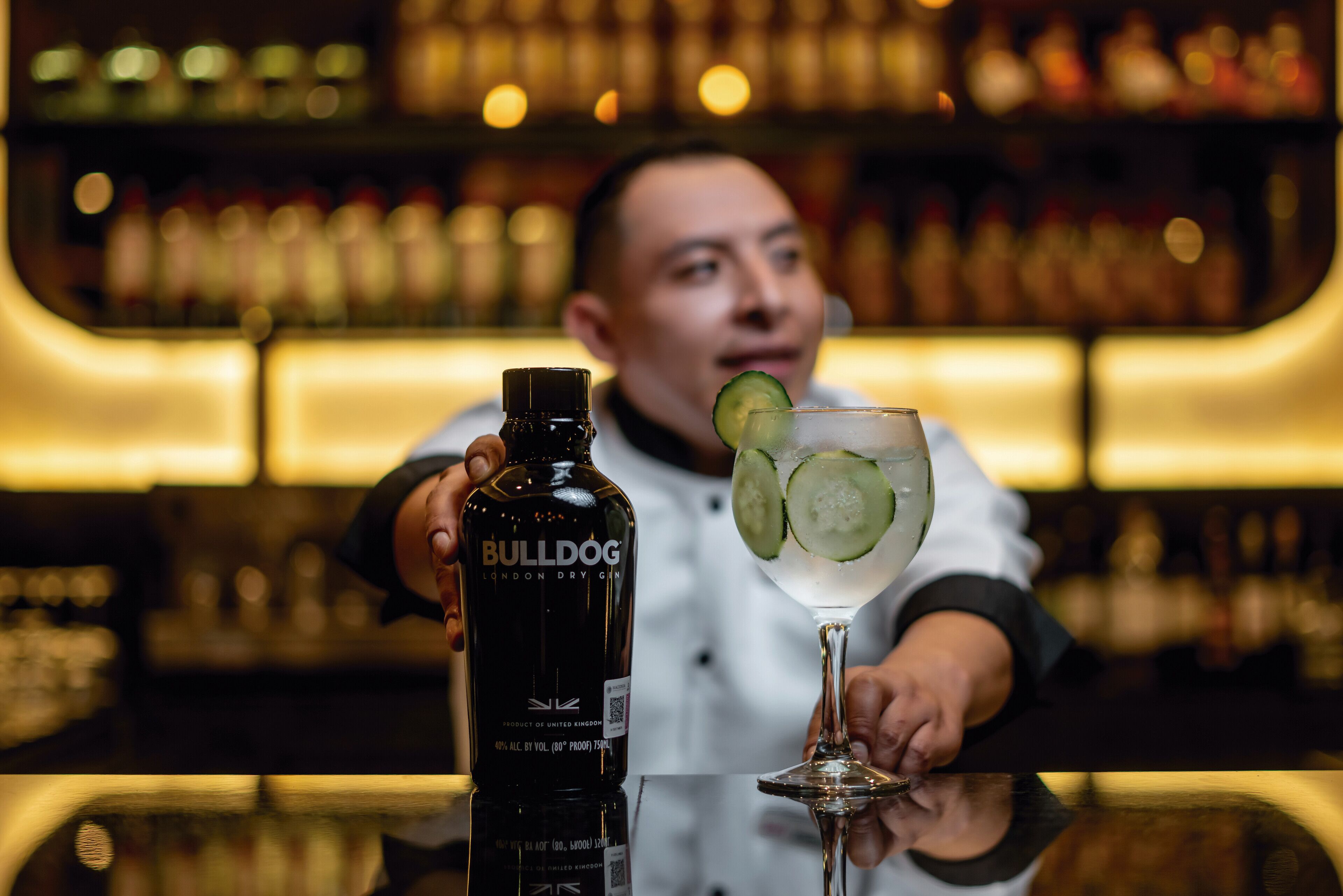 Un barman verse une boisson dans un verre. On aperçoit une bouteille étiquetée « BULLDOG » et, à l’arrière-plan, un comptoir avec des étagères remplies de bouteilles.