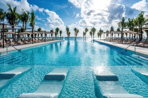 A large swimming pool with clear blue water, surrounded by lounge chairs and palm trees, with a view of the ocean. - Hotel Riu Palace Kukulkan - Adults Only - All Inclusive (Cancun)