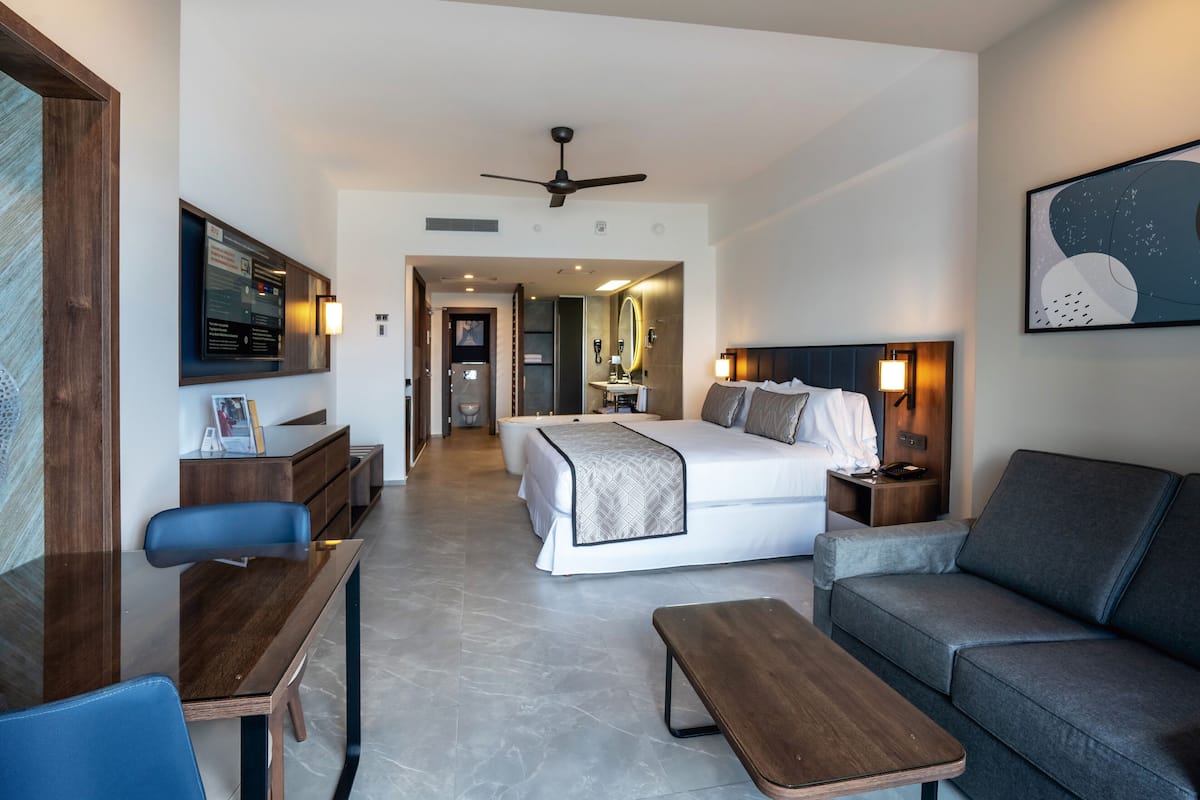 A modern hotel room with a bed, a sofa, a desk, a chair, and a TV.